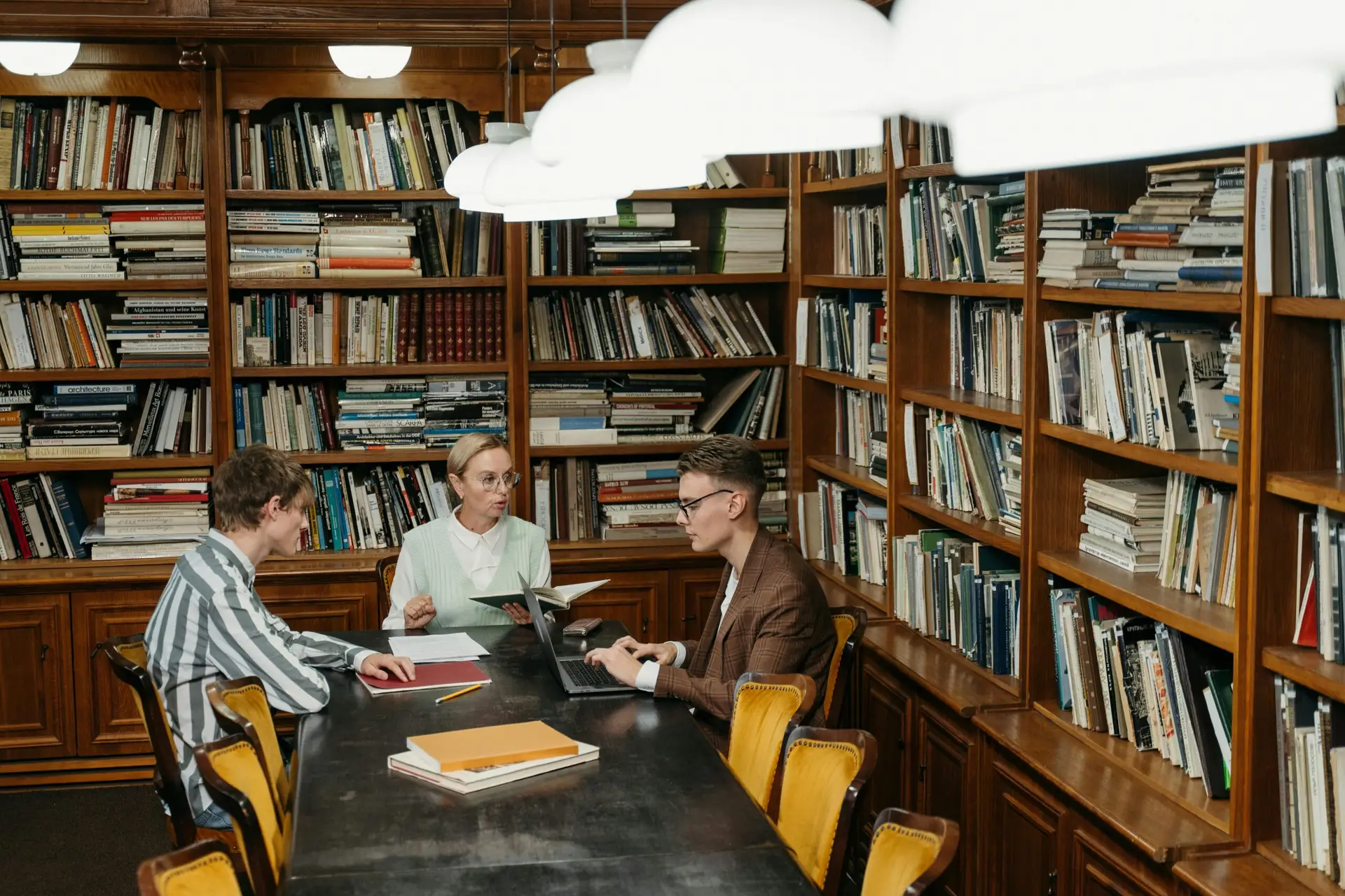 Three professionals having a meeting with laptops and documents in a well-stocked library.
